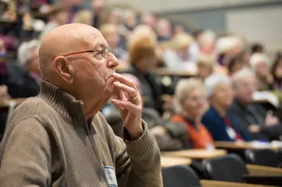 Man sitting in an audience, looking to the right of the image, with his hand at his face, fingers on lips